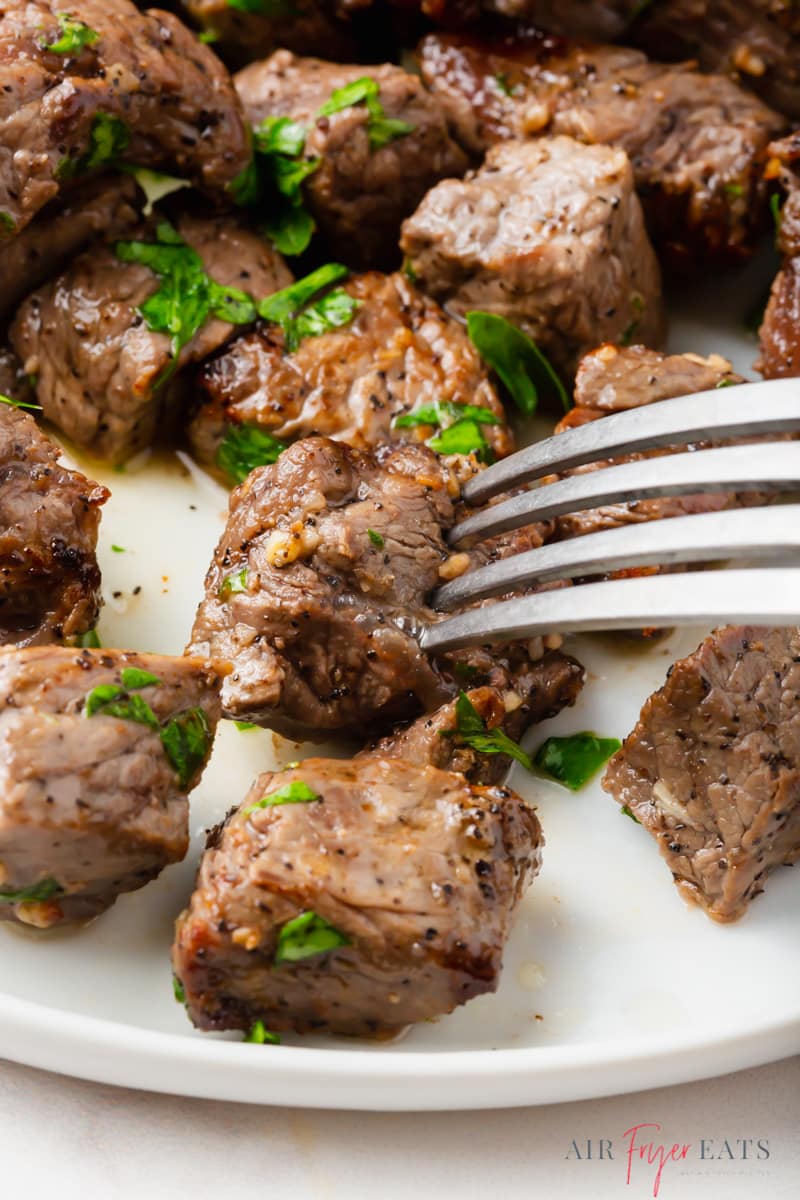 closeup shot of a plate of steak bites topped with parsley, being eaten with a fork.