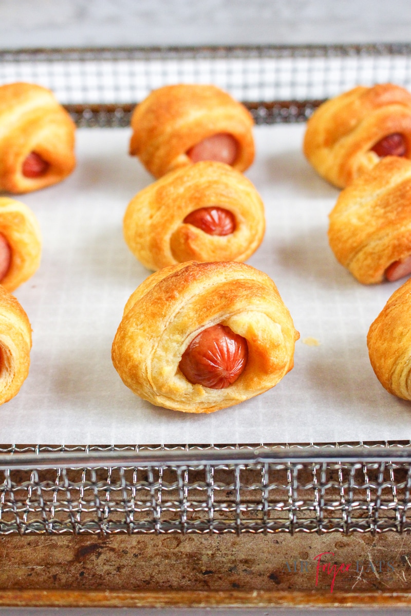 vertical image of a wire rack with white parchment paper and crescent rolls wrapped around hot dogs