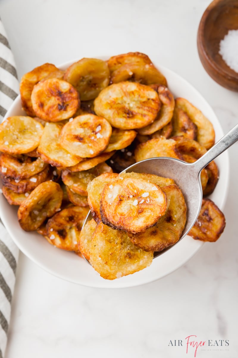 air fryer banana chips in a white bowl. The bowl is sitting on a white marble counter-top and someone is using a silver spoon to scoop a few chips out of the bowl.