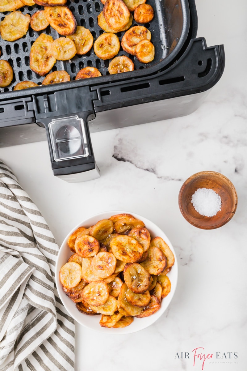 a bowl of banana chips next to an air fryer basket of banana chips on a marble countertop