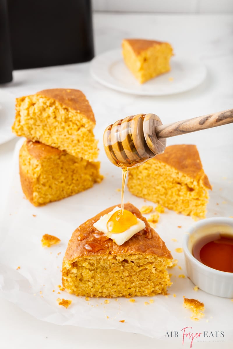 wedges of cornbread on a white platter. One slice has a pat of butter on it and is being drizzled with honey using a honey dipper