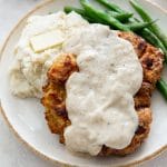 Chicken fried steak with creamy gravy with sides of mashed potatoes and butter and green beans, on a white plate.