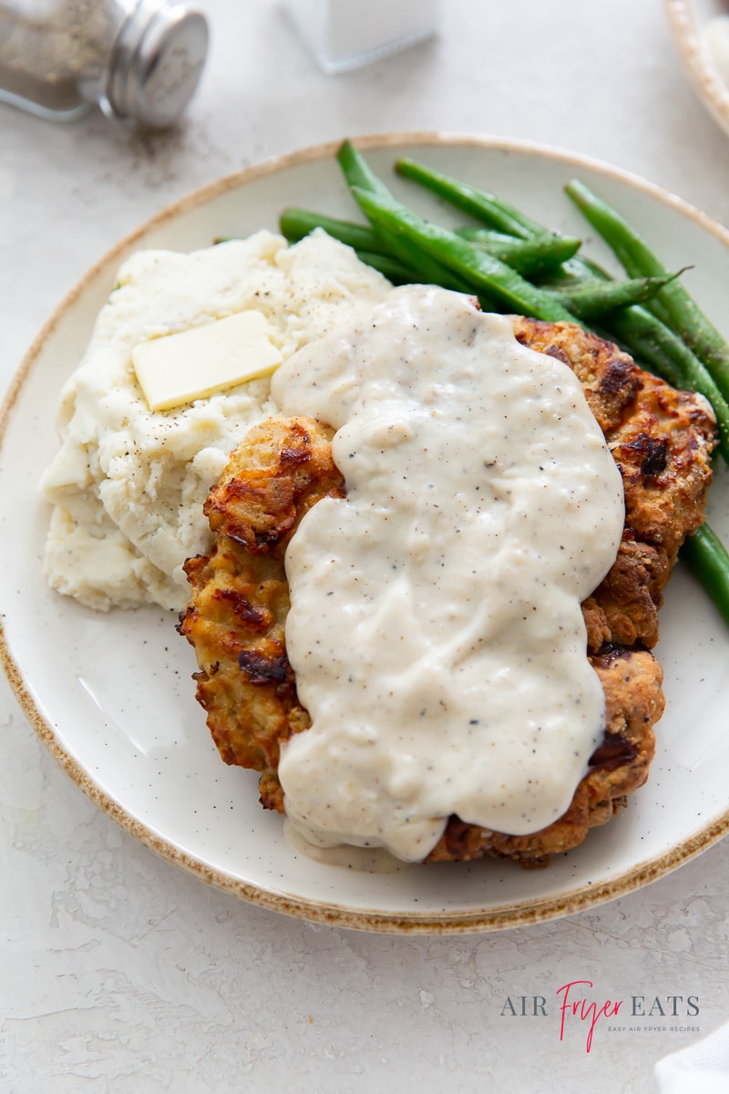 Chicken fried steak with creamy gravy with sides of mashed potatoes and butter and green beans, on a white plate.