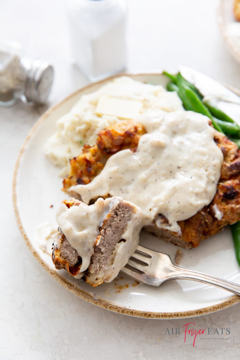 a plate of chicken fried steak with creamy gravy. a slice has been cut and pierced with a fork, showing the inside of the meat, cooked well done.