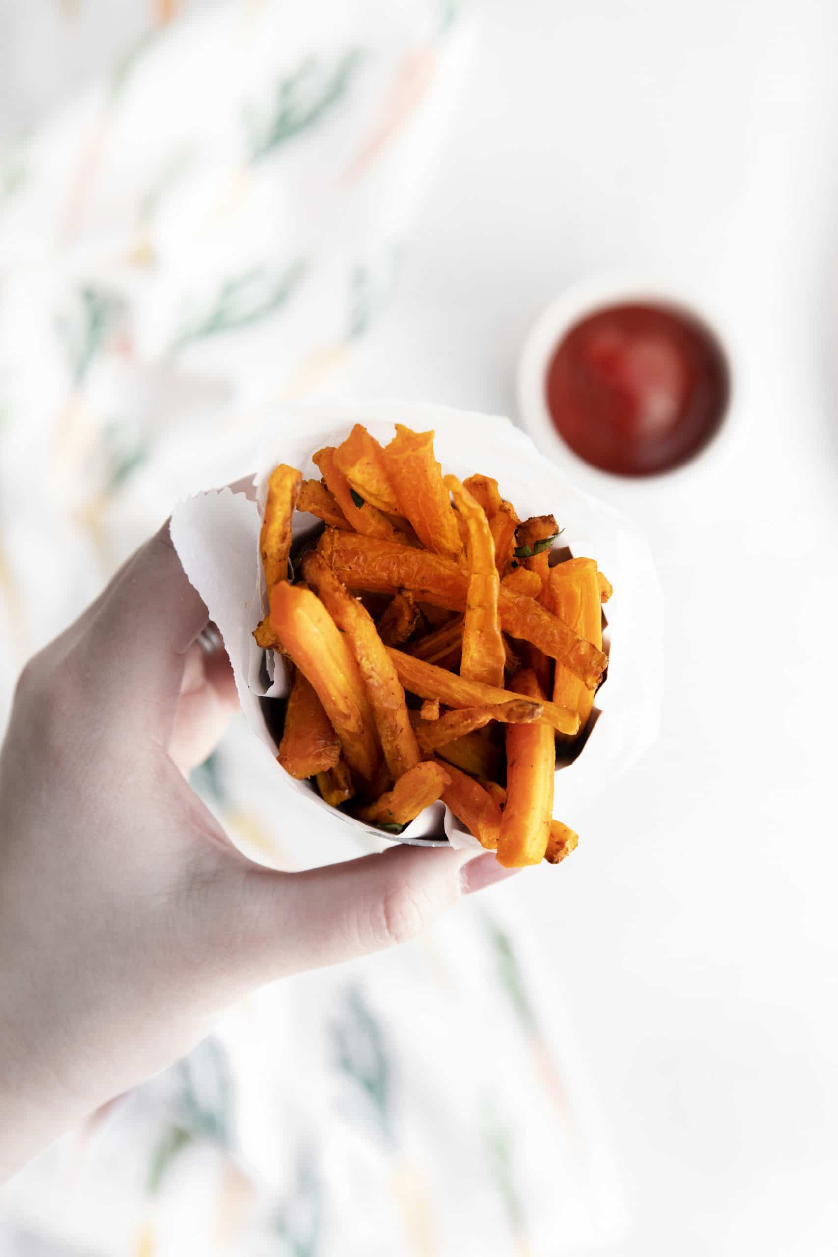a hand holding a cup of air fryer carrot fries. A small cup of ketchup is visible in the background