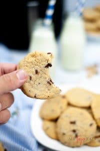 a hand holding a chocolate chip cookie with a bite taken out of it, over a plate of cookies.