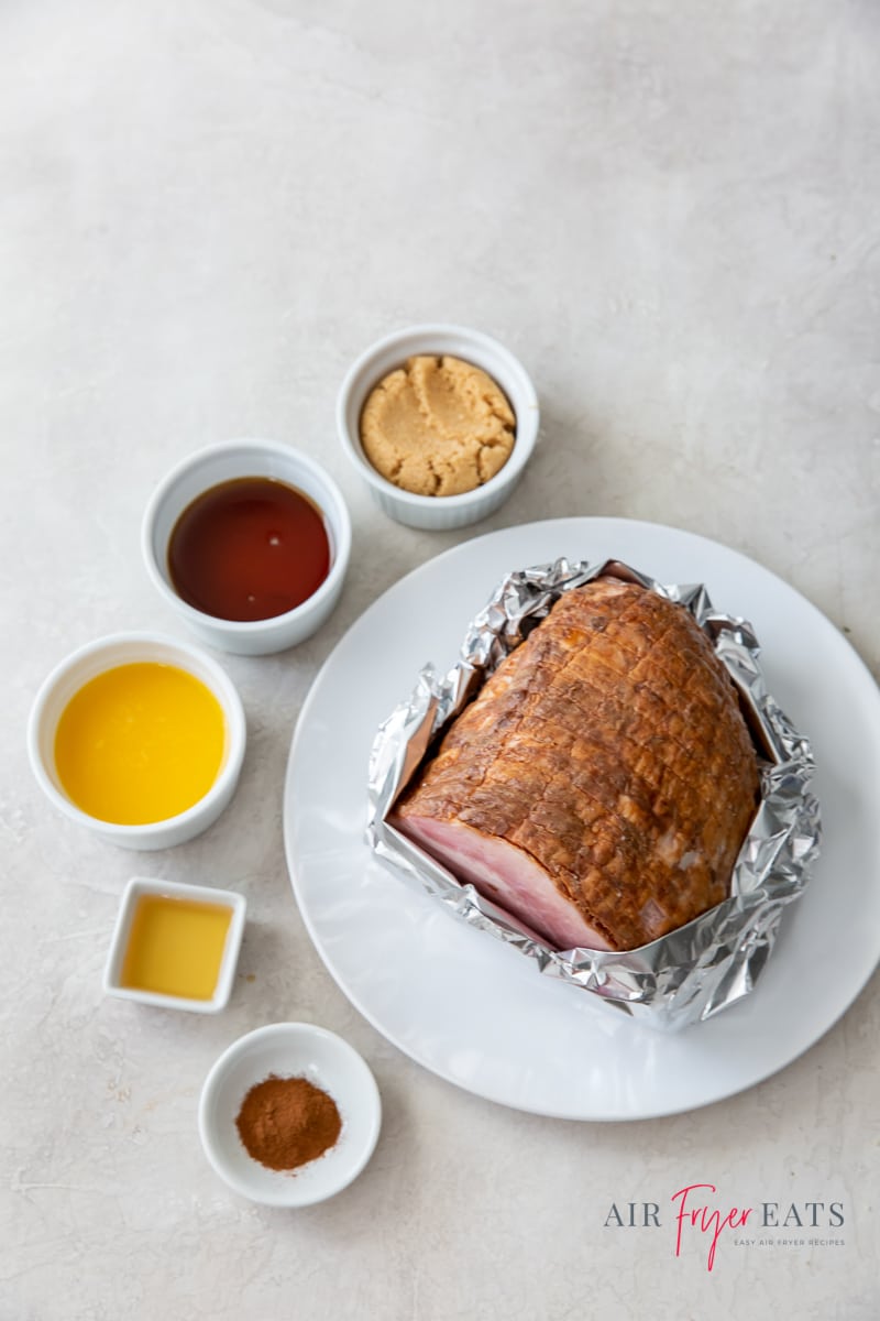a white plate with a ham wrapped in foil on it. To the left are 5 small white bowls with ingredients for ham glaze.