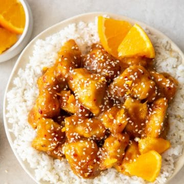 a white plate full of white rice, topped with orange chicken, garnished with orange wedges and sesame seeds