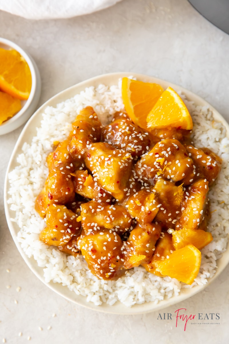 a white plate full of white rice, topped with orange chicken, garnished with orange wedges and sesame seeds