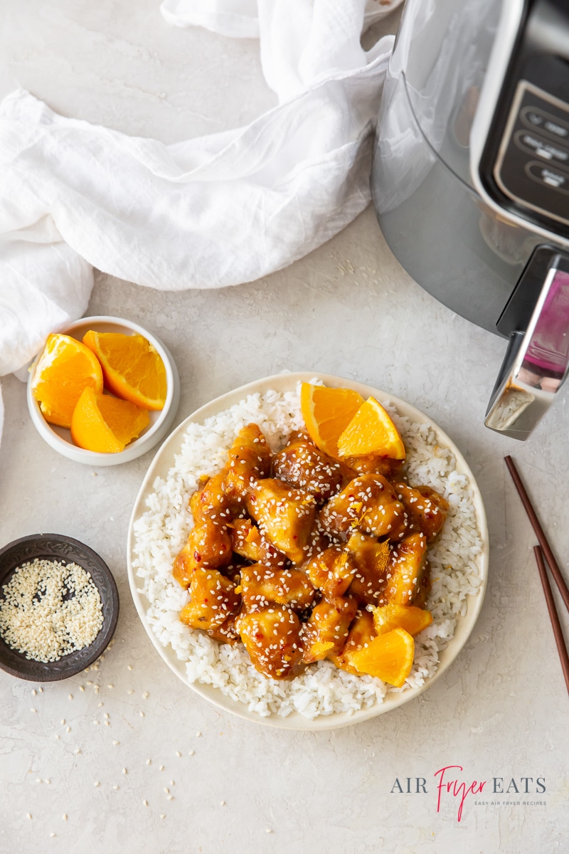 a white plate of rice and orange chicken next to a black air fryer, a bowl of oranges, and a kitchen towel.