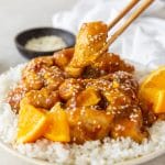 a plate of orange chicken over rice, a pair of chopsticks is grabbing a piece of chicken