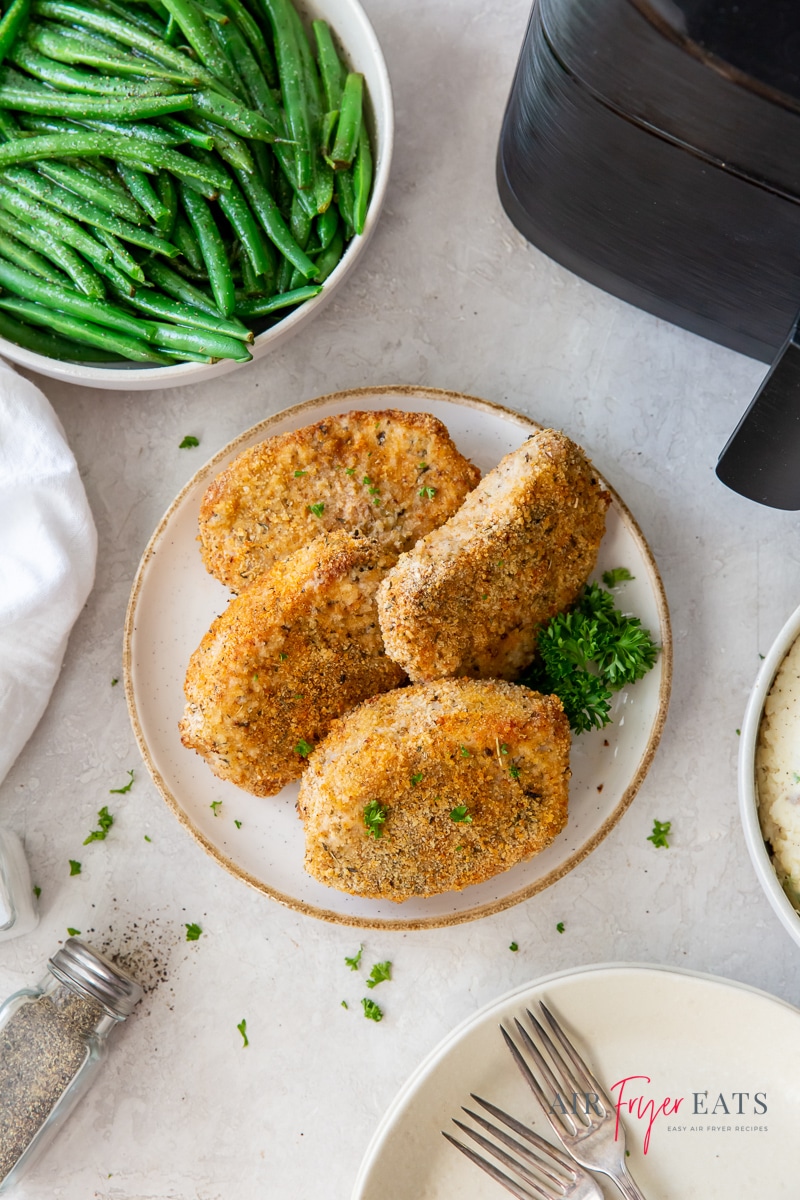 Four breaded pork chops on a round white and tan plate next to an air fryer