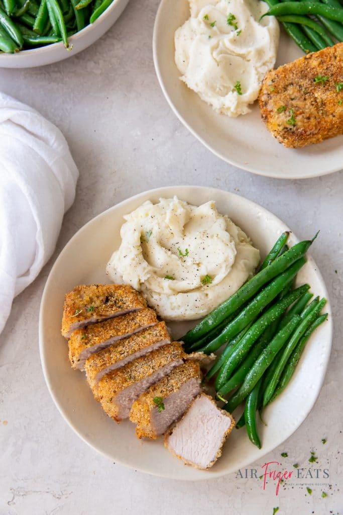 a round white plate with mashed potatoes, whole steamed green beans, and sliced breaded pork chop