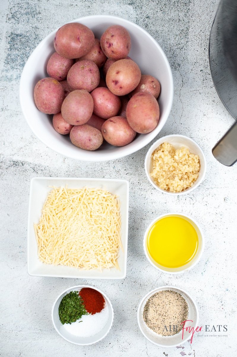 Vertical photo of air fryer roasted red potatoes ingredients. Bowl of red potatoes, minced garlic, Nature's seasoning, olive oil, salt, paprika, parsley and a cup of shredded parmesan