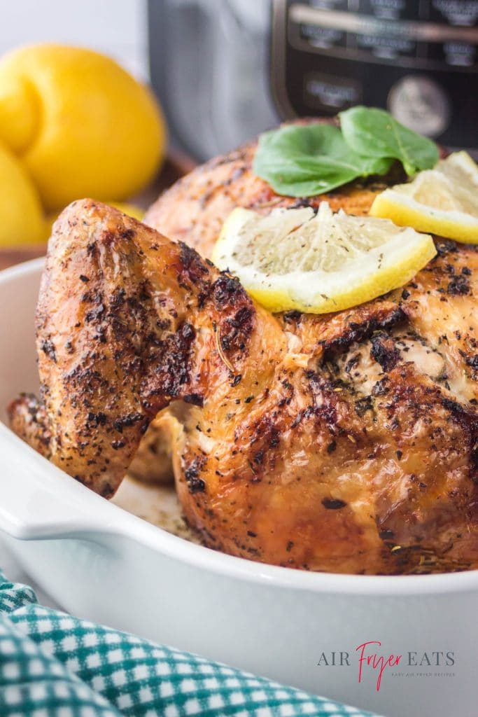 a whole air fried chicken in a white casserole dish, garnished with lemon slices and basil leaves. The chicken is on a counter in front of a ninja foodi air fryer