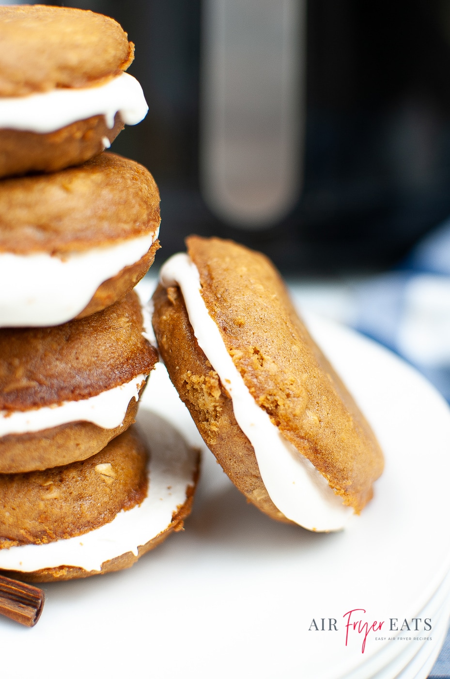 vertical photo of 4 oatmeal creme pies with one pie on the side leaning on the stack of oatmeal pies. White cream is slathered between oatmeal cookies, black air fryer in back with silver handle.