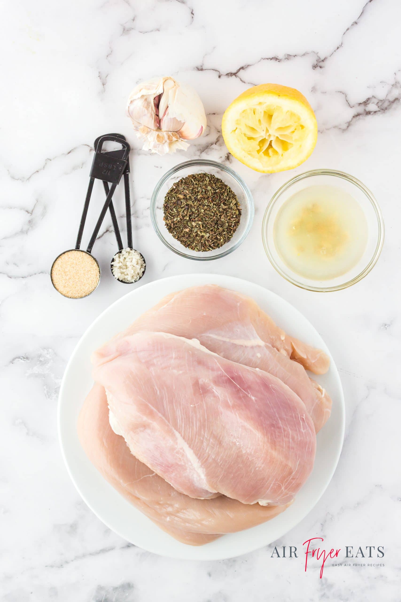 a plate of raw chicken breasts, and small bowls of seasonings, including lemon juice and zest, salt, pepper, basil, and garlic powder.