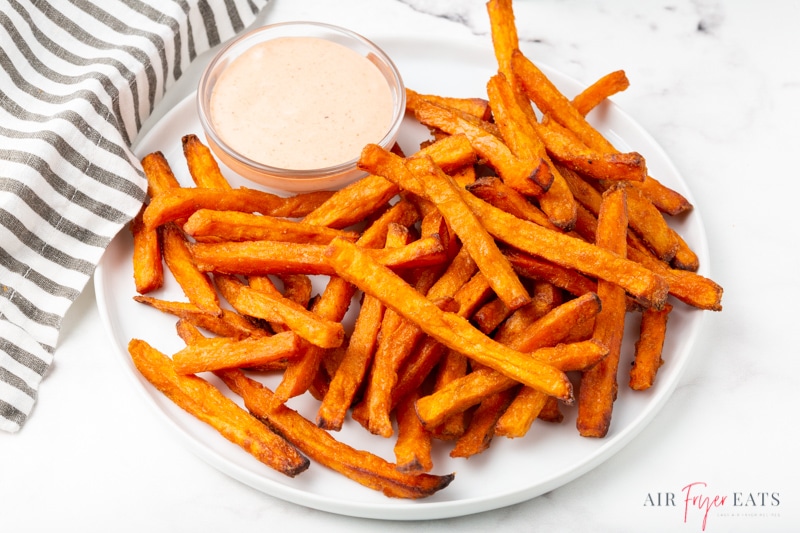 a round white plate filled with crispy air fryer sweet potato fries with a side cup of creamy sauce
