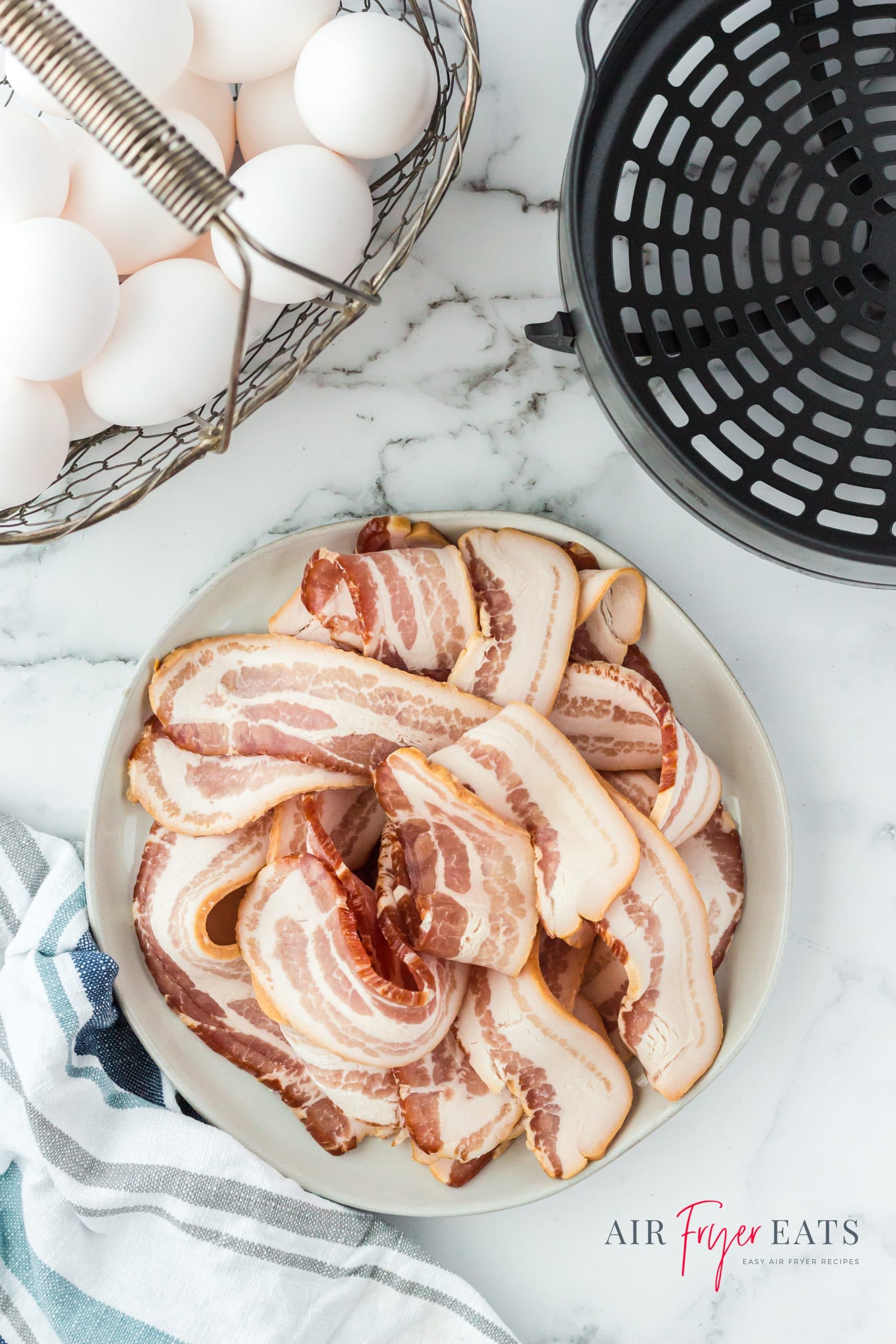 a plate of raw strips of bacon next to a ninja foodi air fryer basket and a basket of eggs.