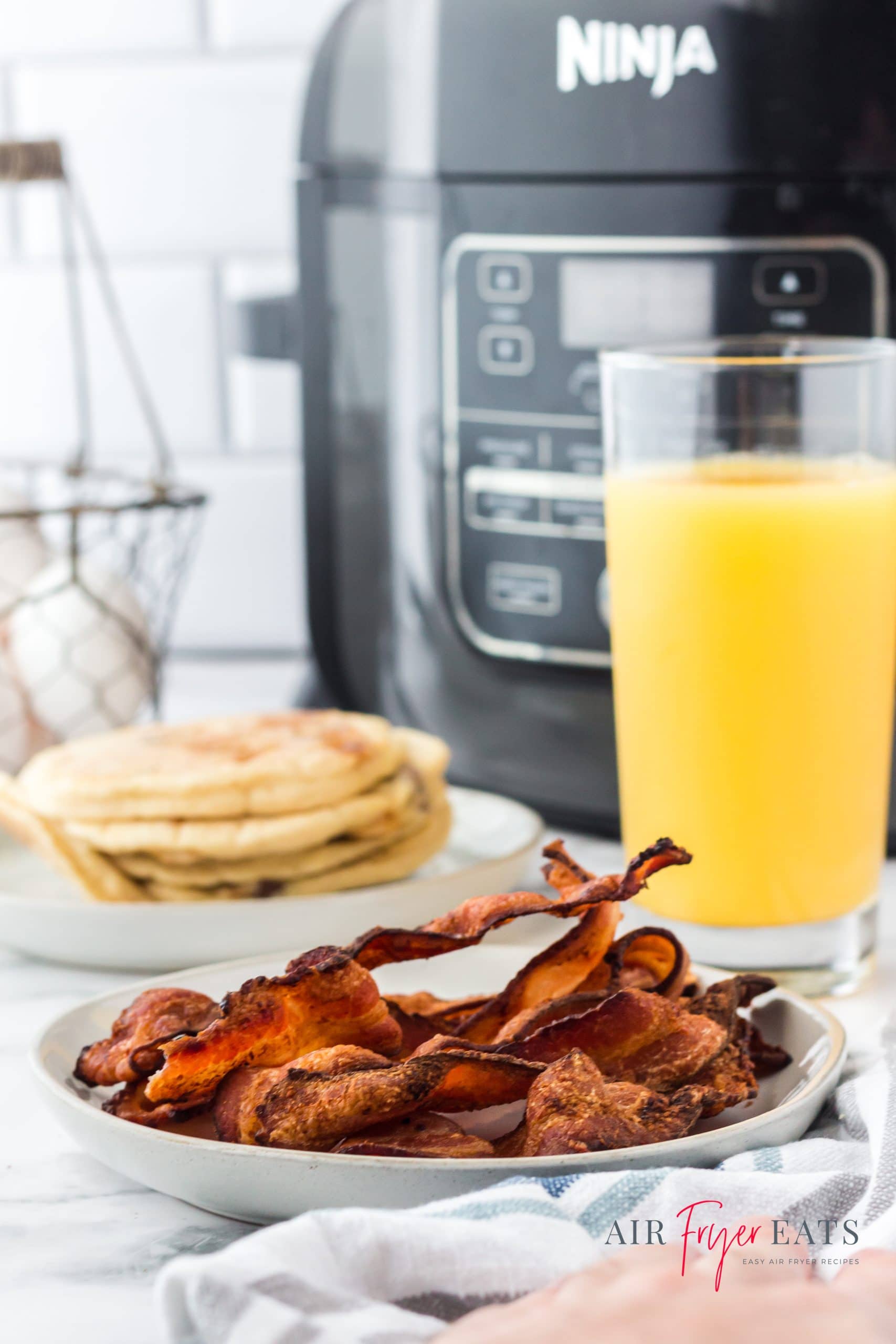 a breakfast of a plate of bacon, a stack of pancakes, and a tall glass of orange juice in front of a ninja foodi multicooker