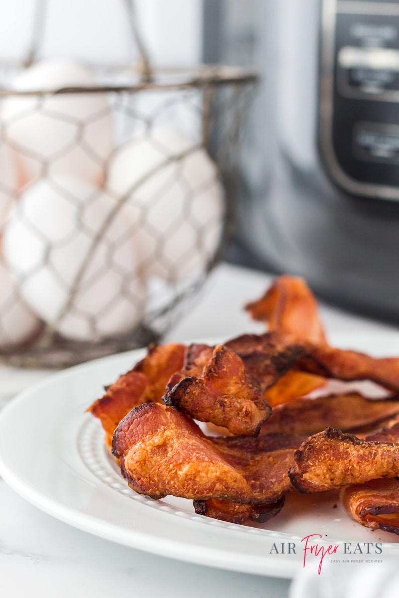 crispy bacon on a white plate in front of a ninja foodi and a basket of eggs