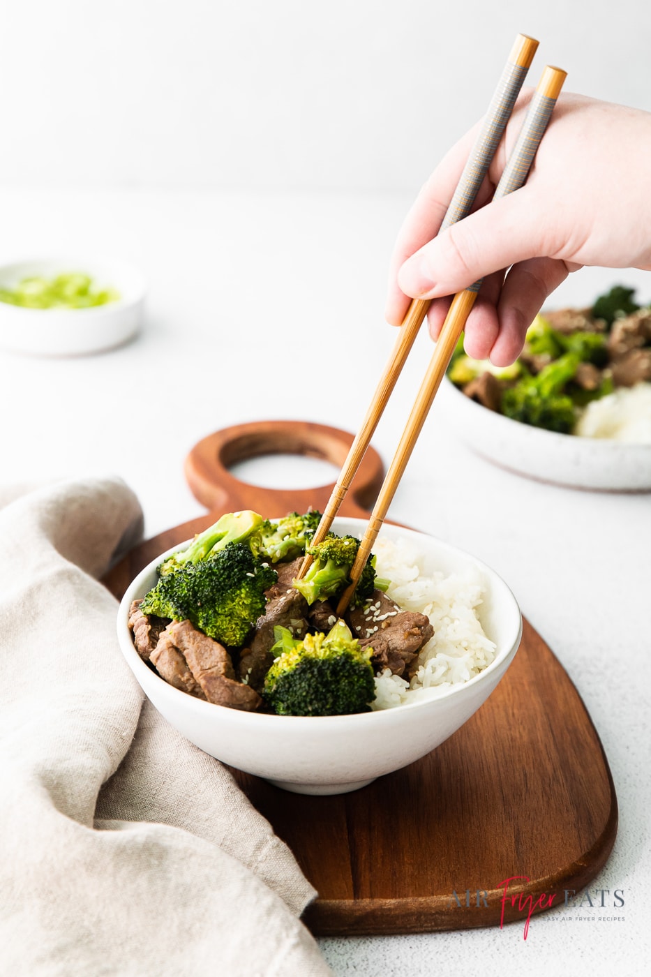 vertical photo of a brown chopping board with a white bowl on containing beef, broccoli and rice. Above the bowl is a hand holding a piece of broccoli between a set of chopsticks
