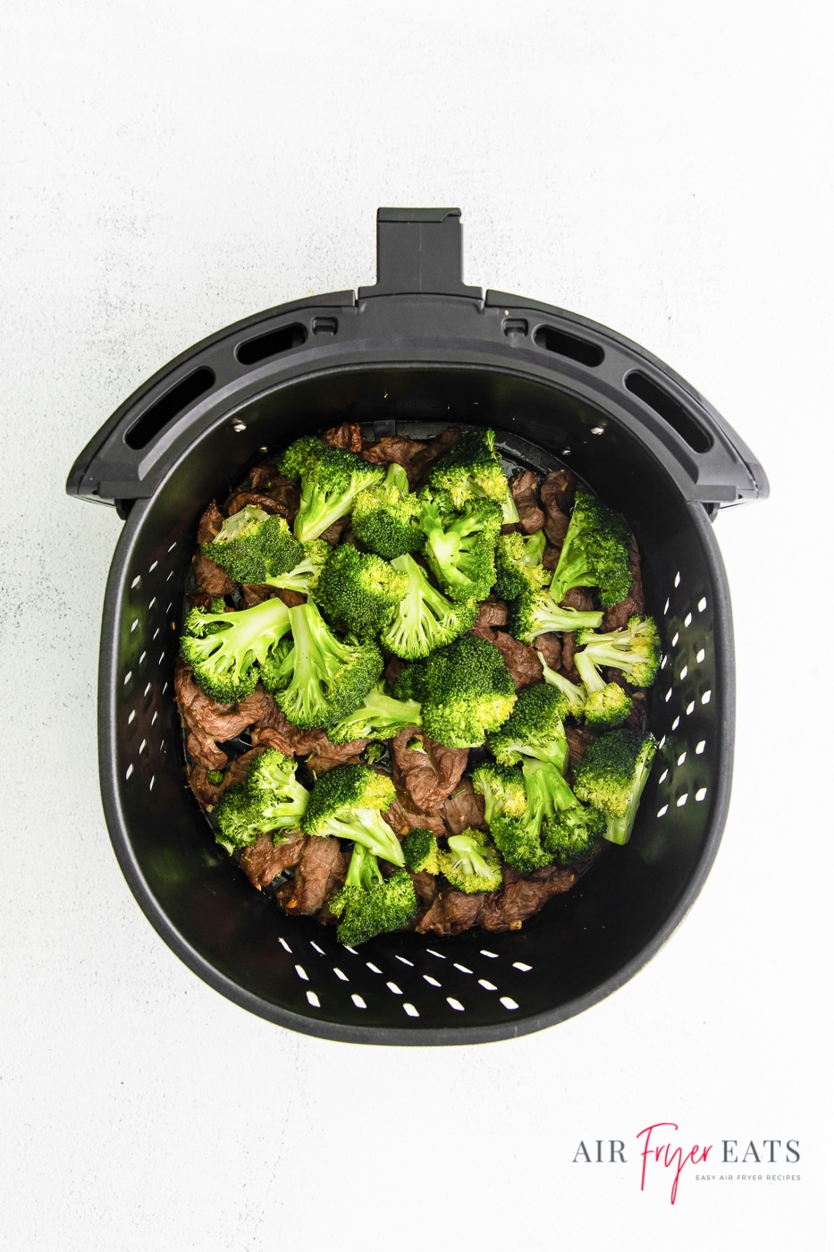 Overhead shot looking down into a black air fryer basket containing cooked beef and broccoli. To the bottom right underneath the basket is the website logo.