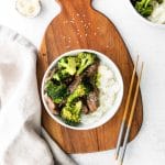 Photo of a white bowl containing beef, broccoli and rice sat on a brown wooden cutting board. Also on the board is a pair of chopsticks. To the left of the board is a white tea towel and below the board is text overlay with the site's logo