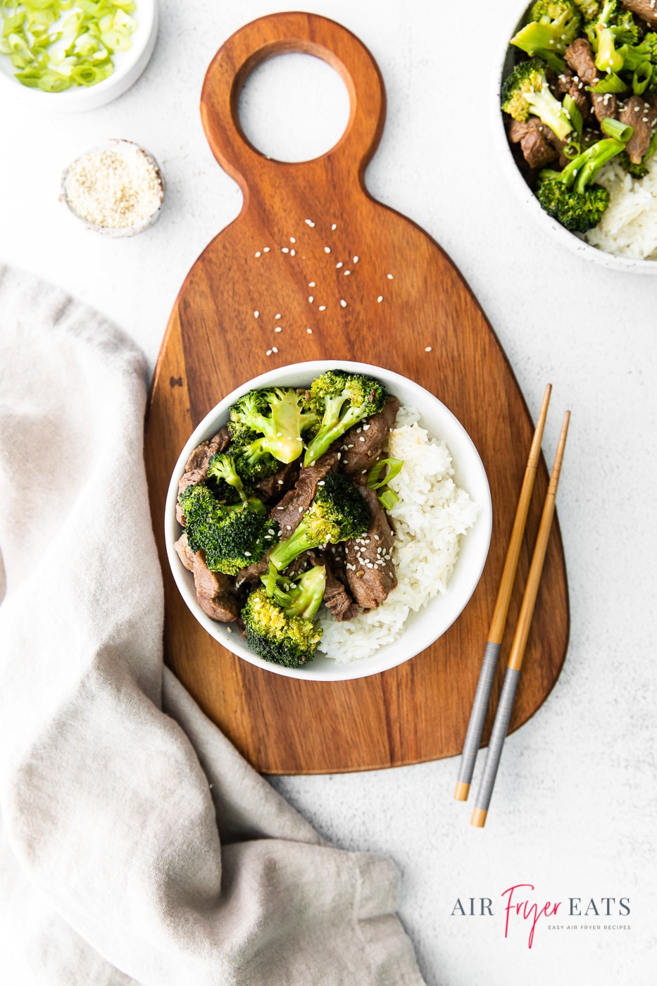 Photo of a white bowl containing beef, broccoli and rice sat on a brown wooden cutting board. Also on the board is a set of chopsticks. To the left of the board is an off-white tea towel and below the board is text with the site's logo