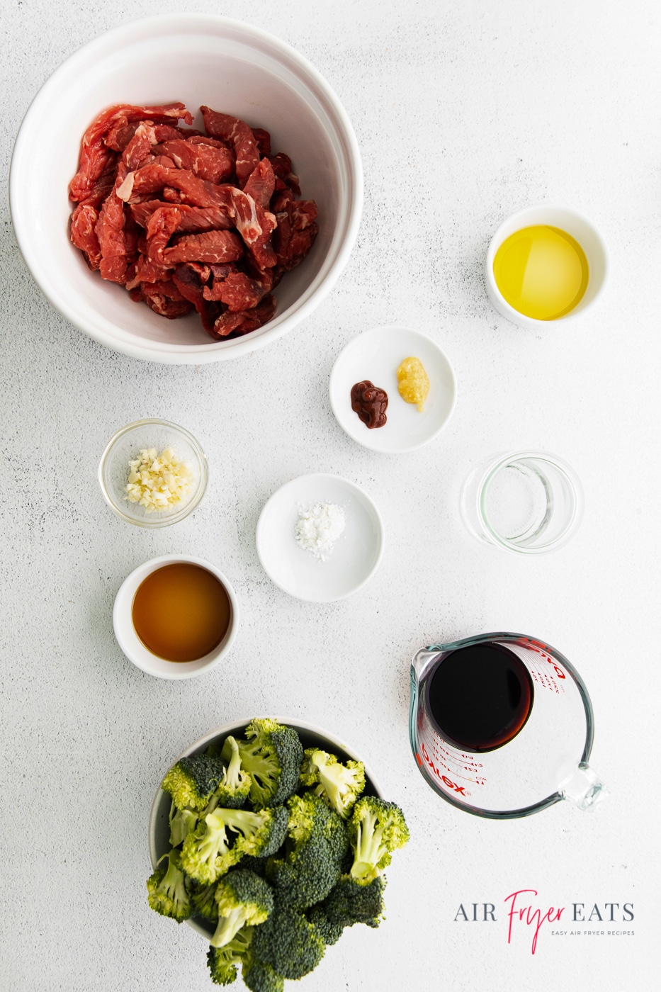 vertical photo of air fryer beef and broccoli ingredients: thinly sliced steak, broccoli florets, soy sauce, water, sesame oil, minced garlic, grated ginger, sriracha, olive oil and cornstarch