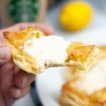vertical photo of a hand holding an air fryer Starbucks cream cheese danish with a bite taken from it. Background right is a plate with more air fryer Starbucks cream cheese danish. Slightly further back is a clear plastic Starbucks cup containing light brown liquid with a lemon to the right of it