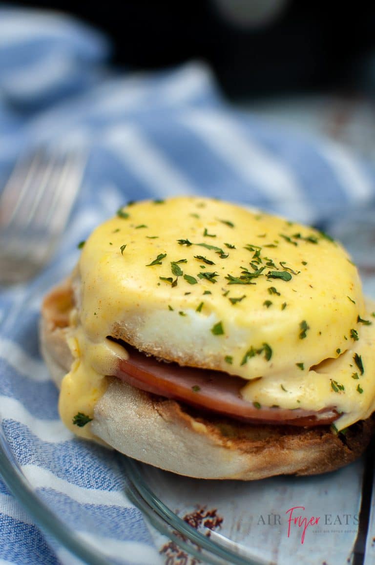 Close up image of a single air fried eggs benedict sat on a glass plate. The plate is on top of a blue and white tea towel with a fork just to the left hand side