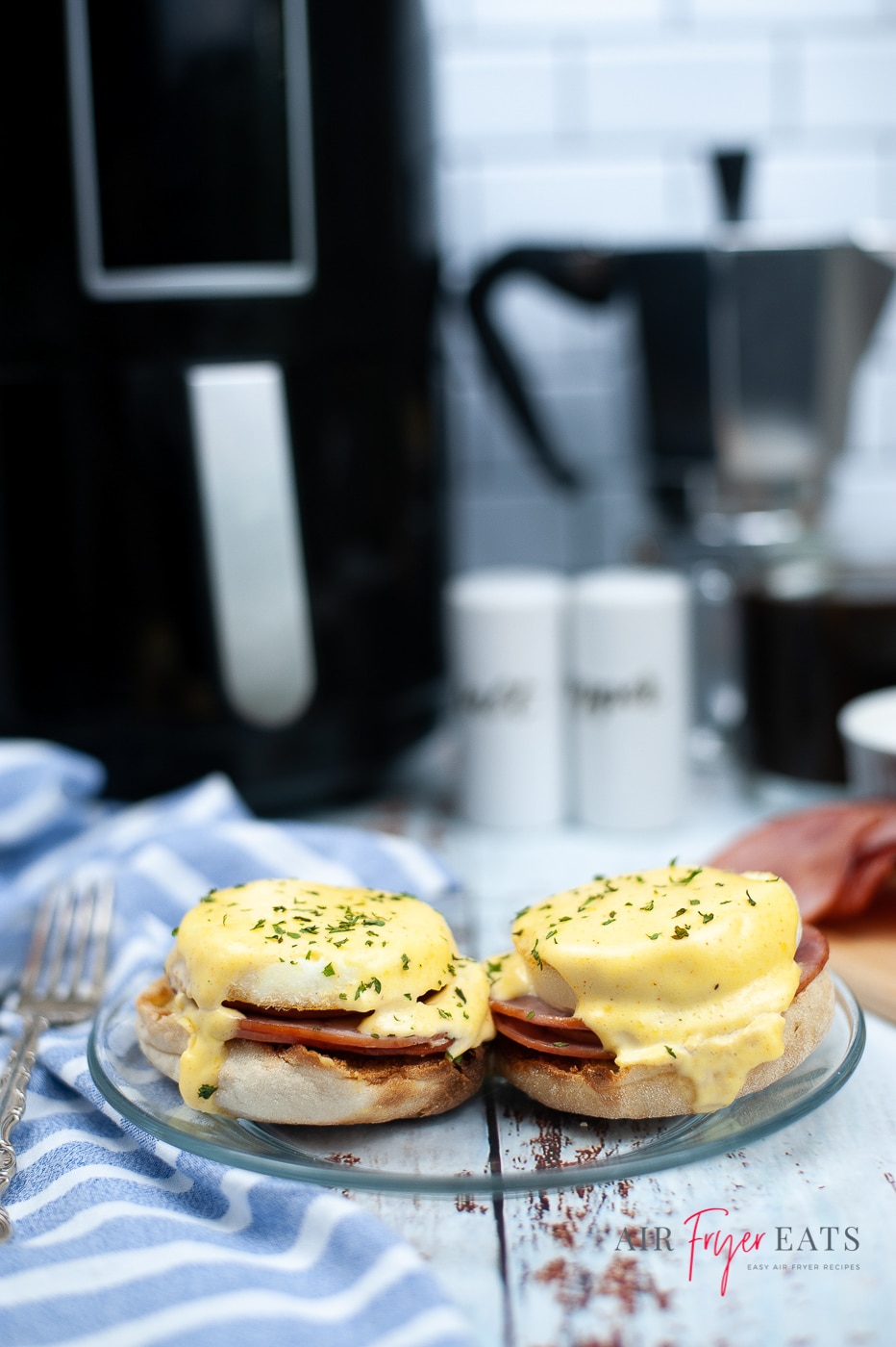 vertical photo of two egg benedict sandwiches on a glass plate. Toasted English muffin halves topped with air fried Canadian bacon with soft eggs with a yellow hollandaise sauce poured over. In the background is a black air fryer with silver handle. Below the plate is the website name and logo.