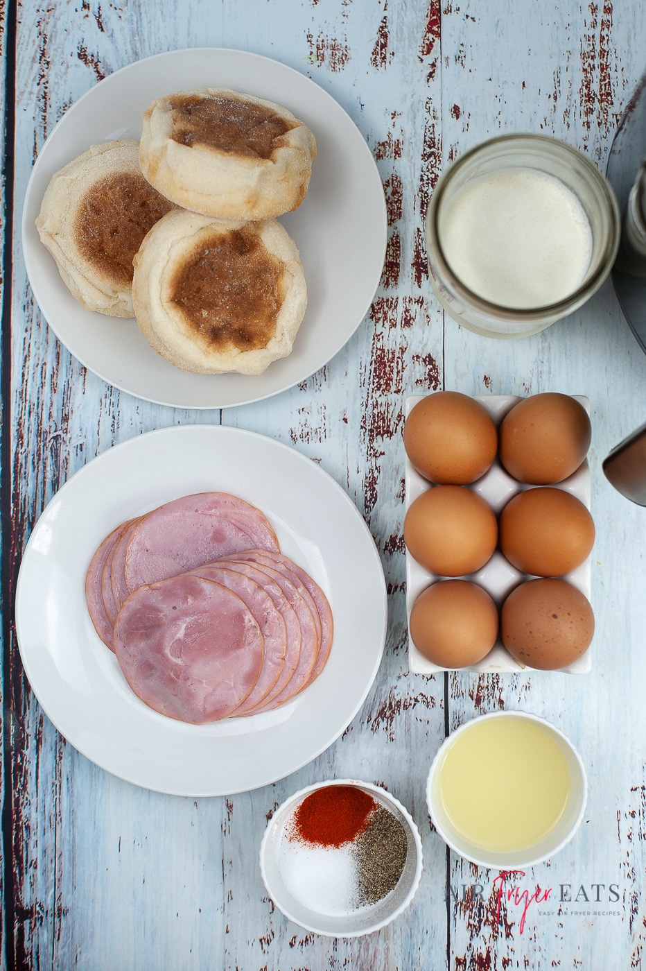 Overhead shot of ingredients for air fryer eggs benedict. 3 English muffins, sliced Canadian bacon, bowl of spices, lemon juice, eggs and cream