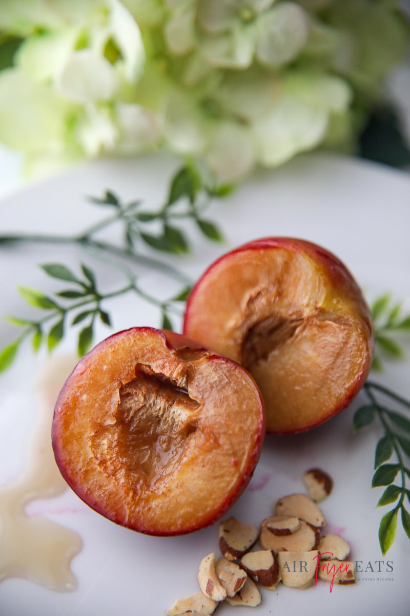 Vertical photo of two air fried plum halves on a white plate drizzled with syrup. Also on the plate are some chopped nuts and green leaves for decoration. In the background are some white flowers.