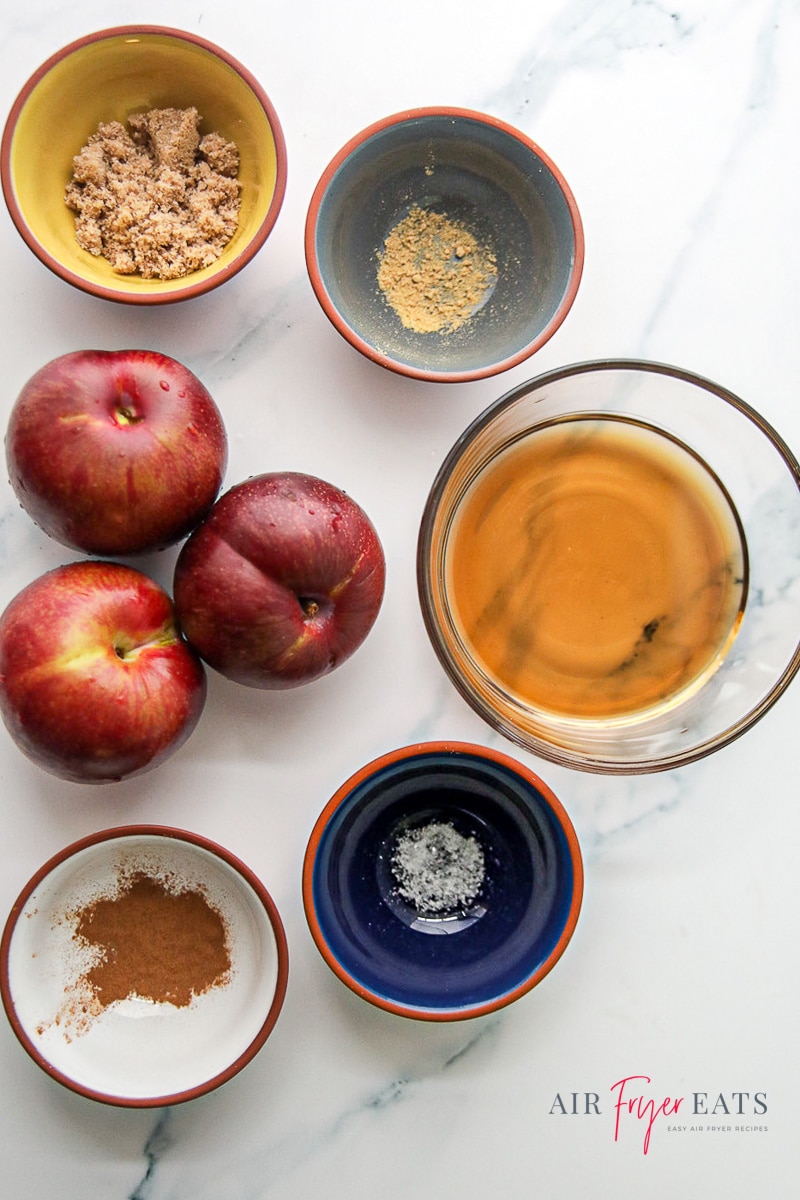 Vertical photo of ingredients. 5 bowls surrounding 3 plums. Each bowl contains an ingredient of brown sugar, ground ginger, ground cinnamon, salt and maple syrup.