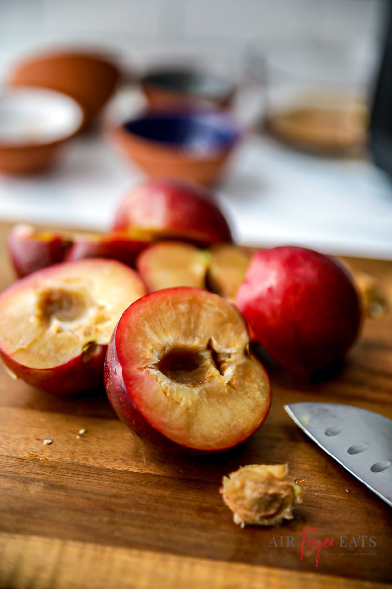 Vertical photo of a close up wooden chopping board with halved plums and a kitchen knife. In the background, there are bowls that have the recipe ingredients in.