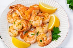 Cooked golden orange shrimp on a white plate. Three lemon quarters on plate with a green herb garnish. The photo is close range.