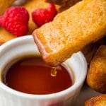 a cup of maple syrup next to cooked french toast sticks and mixed berries