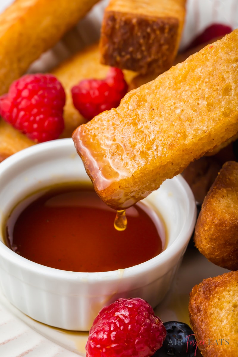 a cup of maple syrup next to cooked french toast sticks and mixed berries