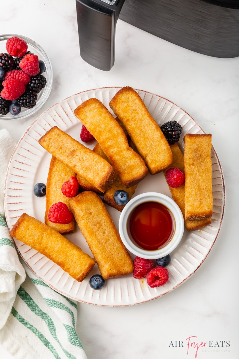 a round white plate filled with french toast sticks and fresh raspberries and blueberries, plus a small cup of maple syrup