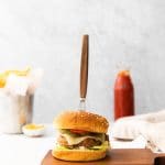 Vertical photo of a brown chopping board with an air fried turkey burger sat on top with a knife stabbed lengthways through the top of the burger.. To the left of the board is a knife on a white table. At the bottom of the image is the Air Fryer Eats logo