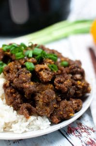 Close up vertical photo of air fried orange beef with rice on a white plate. In the background there is a black air fryer
