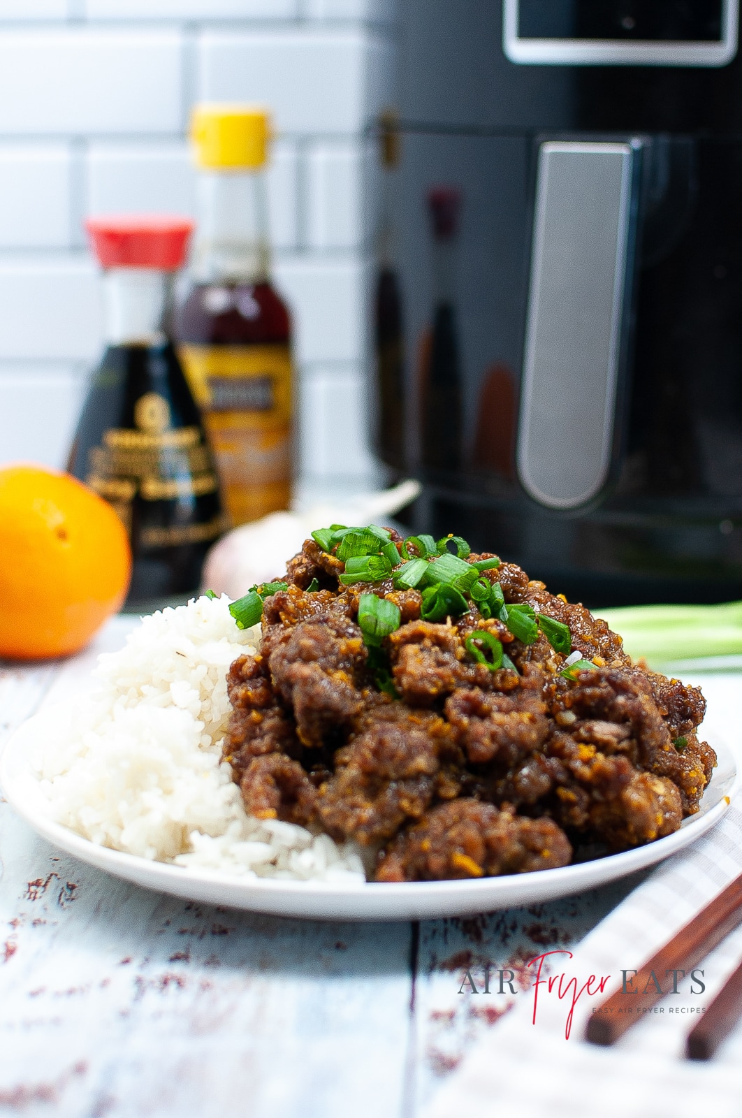 Vertical photo of a plate containing air fried orange beef & rice with green onions on top. In the bacground there is a black air fryer with silver handle. Also in the background are two bottled sauces and an orange.