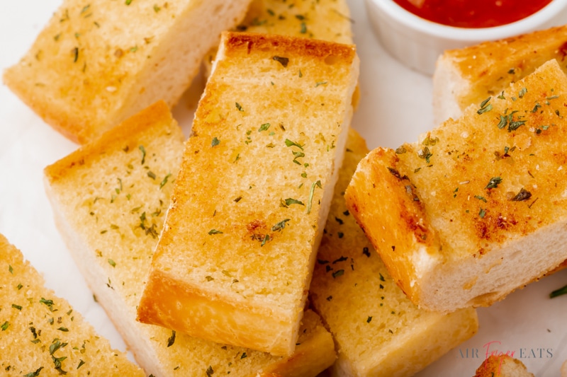 horizontal image, closeup view of slices of garlic bread on a plate next to a cup of marinara sauce. 
