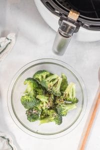 vertical overhead shot of cut broccoli florets in a glass bowl, white air fryer with basket at top right and wooden spoon to right of bowl