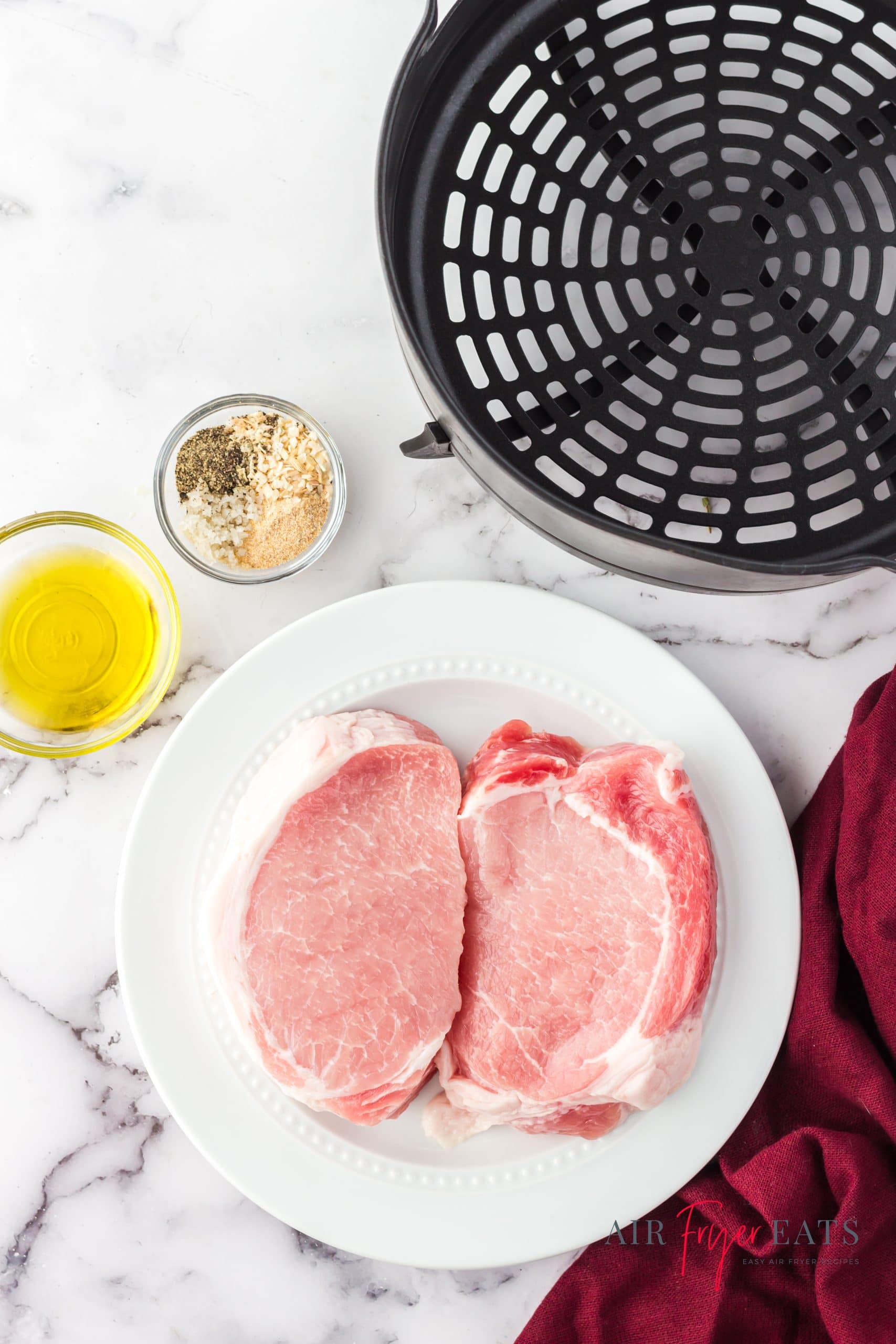 The ingredients for making pork chops in the ninja foodi, including two raw pork chops, oil, seasonings, and the ninja foodi air fryer basket