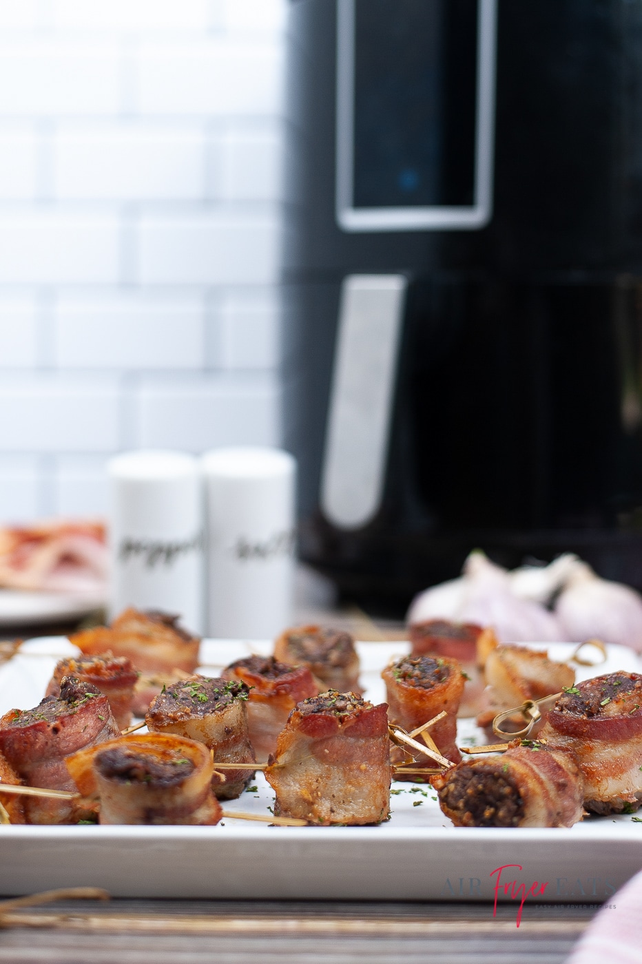 Vertical photo of a white plate containing bacon wrapped steak bites. In the background there is a black air fryer with silver handle. To the left of the air fryer is a salt tub and a pepper tub.
