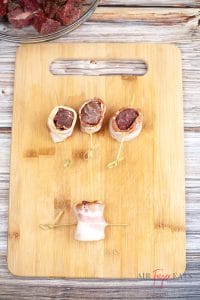 Light brown wooden chopping board with four bacon wrapped steak bites on