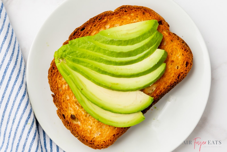 a slice of toast on a small. white plate. The toast is topped with a fan of sliced avocado